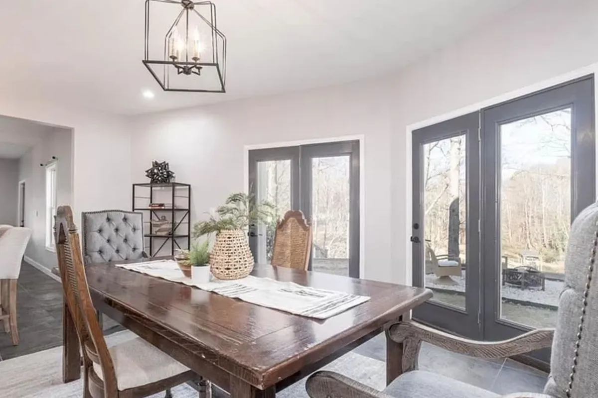 A warm, inviting interior dining area featuring a wooden table adorned with a decorative lantern and greenery in a vase. Large windows let in ample natural light, suggesting a welcoming space for family meals or casual gatherings.