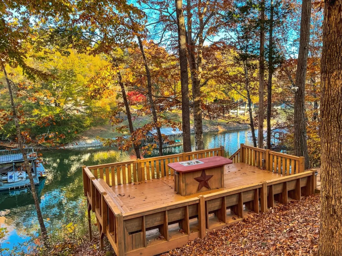 A spacious wooden deck with a clear view of the water, framed by autumn foliage. The deck features ample space for gatherings and leisure, with a distinctive star motif and cozy seating areas, surrounded by lush greenery and a tranquil lakeside setting.
