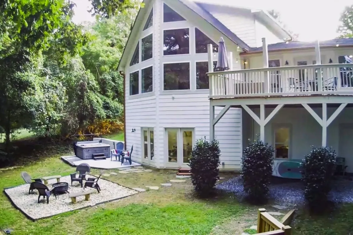 An exterior deck area featuring a hot tub, fire pit surrounded by cozy seating, large windows revealing the home's interior, and a beautifully landscaped yard. The deck overlooks scenic water views, offering a seamless connection between indoor and outdoor living.