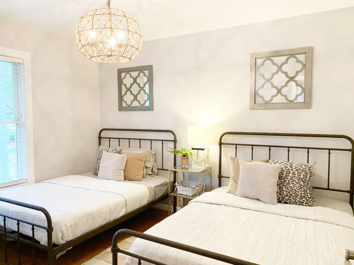 A welcoming bedroom with two metal-framed beds, decorative wall art, a table lamp with a potted plant, and cozy bedding. The space exudes comfort and flexibility, suited for families or groups.