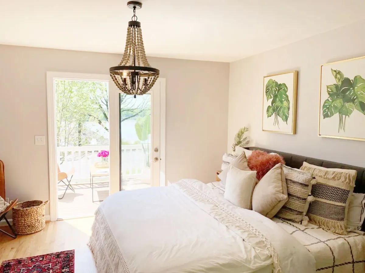 A serene bedroom with a comfortable bed, chandelier overhead, sliding glass doors leading to an outdoor area, and calming plant artwork. Soft natural light fills the room, creating a peaceful atmosphere ideal for relaxation and privacy.