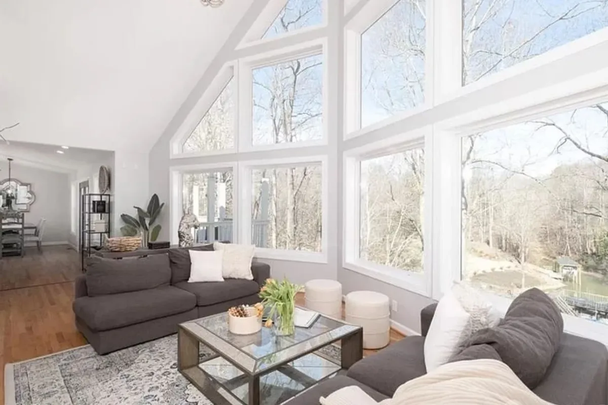 The image showcases a warm and inviting living room with plush, comfortable seating arranged around a stylish coffee table. The space is bathed in natural light streaming through expansive windows that offer breathtaking views of Lake Norman, seamlessly blending the beauty of the outdoors with the cozy interior. The setting is perfect for relaxing, socializing, or simply soaking in the serene lakefront ambiance.