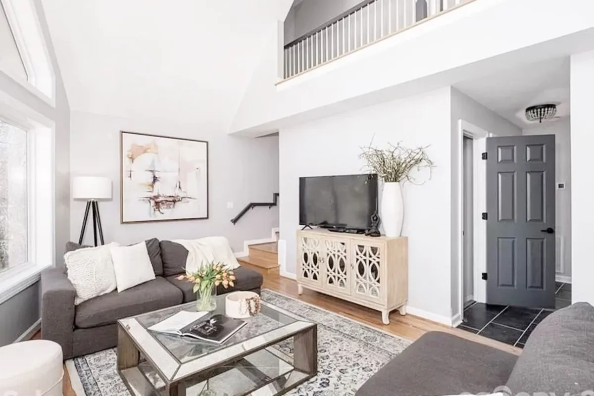 A spacious and inviting living room featuring a comfortable sofa centered around a coffee table, vibrant artwork on the walls, and a large television. Natural light pours in from surrounding windows, creating a cozy and communal atmosphere ideal for gatherings and relaxed movie nights.