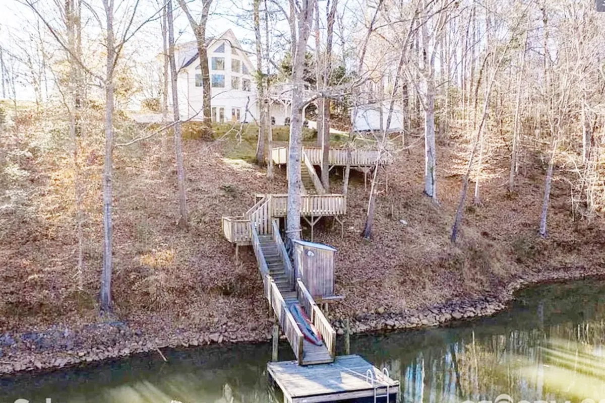 A house perched on a hill overlooks Lake Norman, with wooden stairs leading down through trees to a private dock on the water. The scene is framed by lush greenery and an inviting lakeside atmosphere.