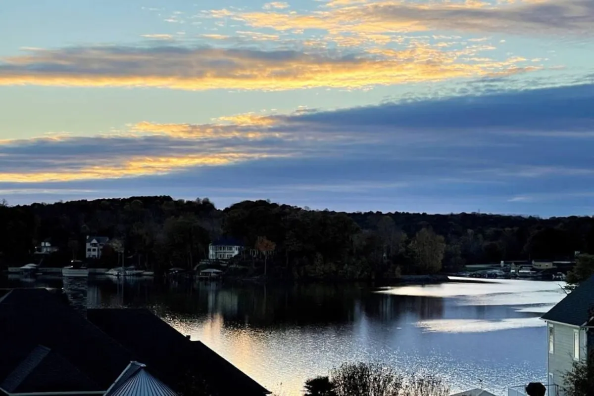 A panoramic view of the calm lake at sunset, surrounded by trees and distant houses. The serene sky and reflective water evoke an inviting atmosphere for lakeside walks and exploration, highlighting the natural beauty of the area.