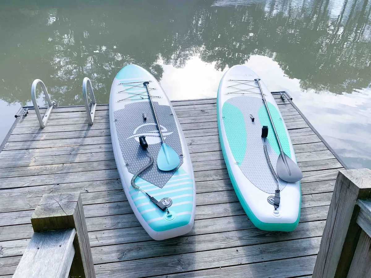 A serene lakeside scene focused on a wooden dock with two stand-up paddleboards resting on it, reflections glimmering on the water. The image evokes water-based adventure and easy access to lake activities.