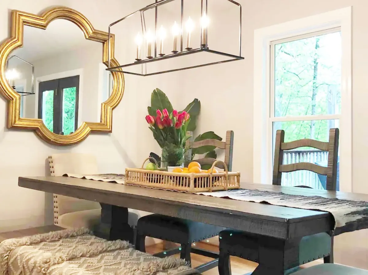 A beautifully set dining table beneath a glowing chandelier, with an arrangement of flowers and a mirror reflecting warm light. Large windows offer a view, enhancing the space for both formal dinners and casual brunches.