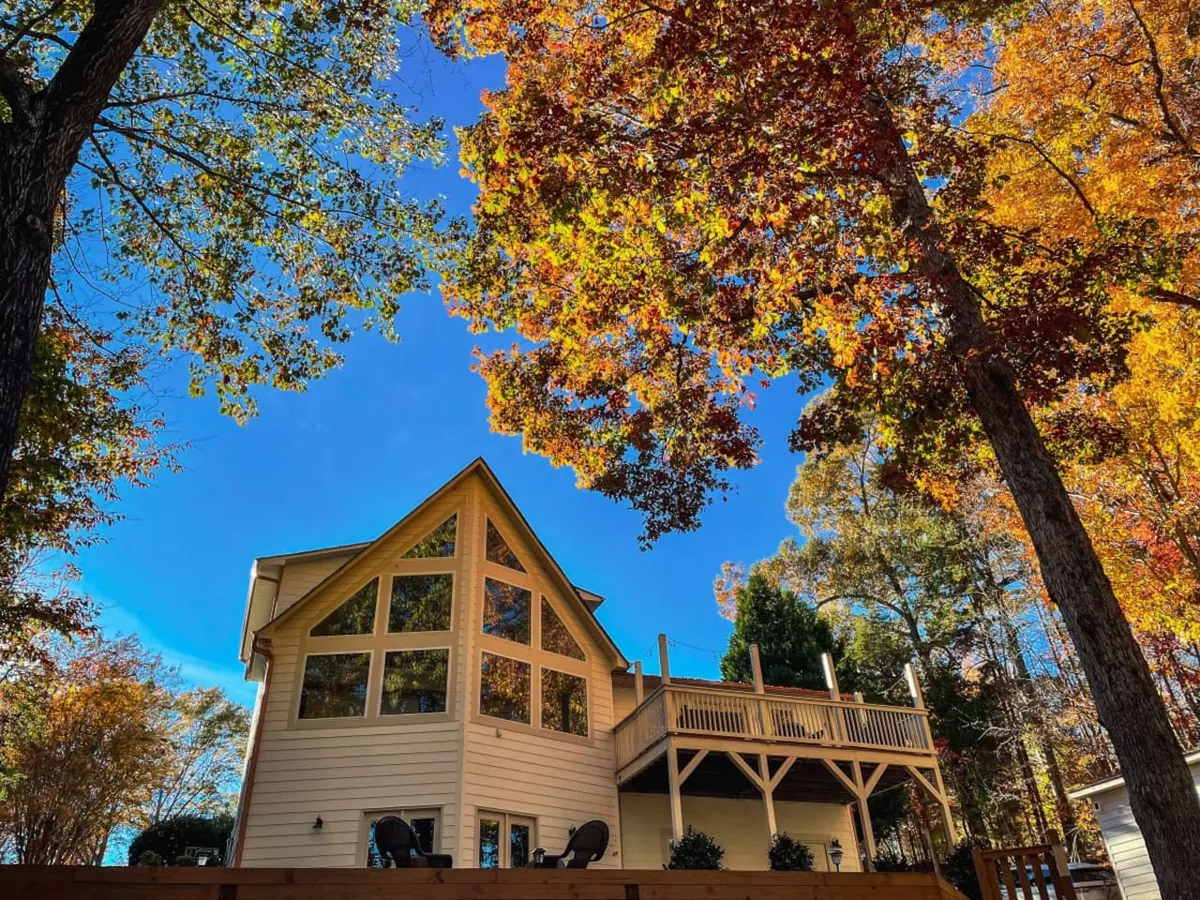 The house with expansive windows and a large wooden deck, surrounded by vibrant, colorful autumn trees. The deck provides space for outdoor relaxation and socializing, while the house sits cozy in a quiet, scenic neighborhood setting.
